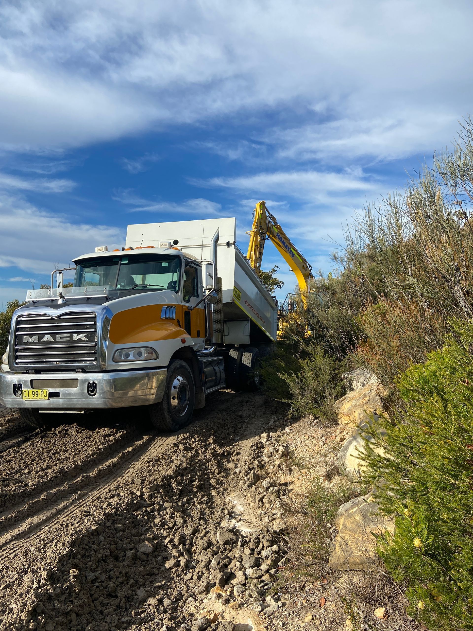 A dump truck is driving down a dirt road next to a yellow excavator. — Adam Lowe Earthmoving Pty Ltd in Somersby, NSW