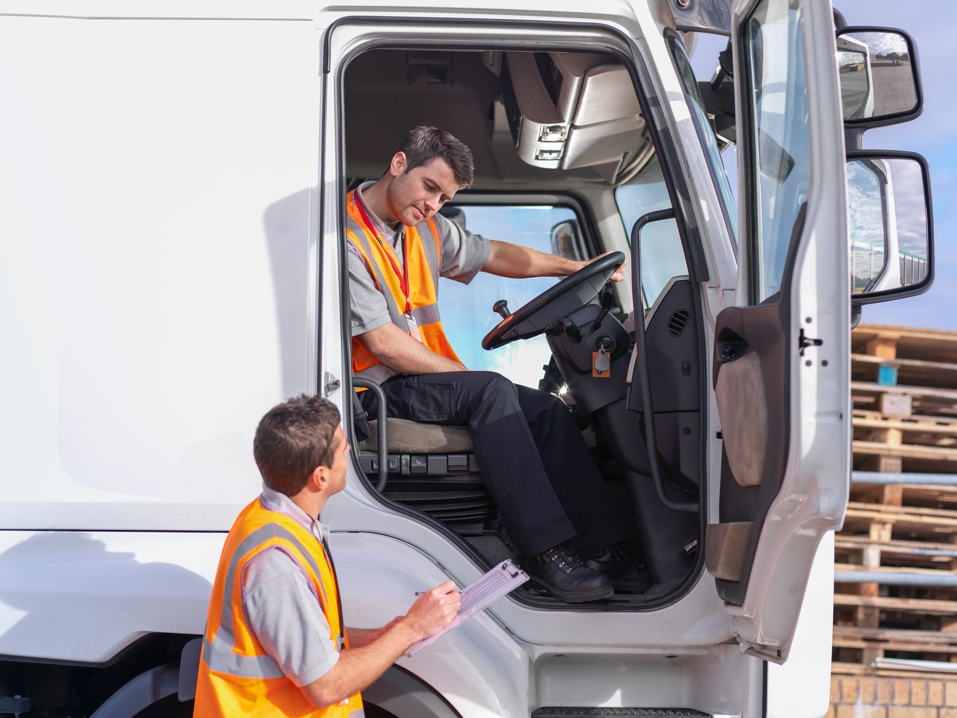 A man is sitting in the driver's seat of a truck while another man writes on a clipboard.