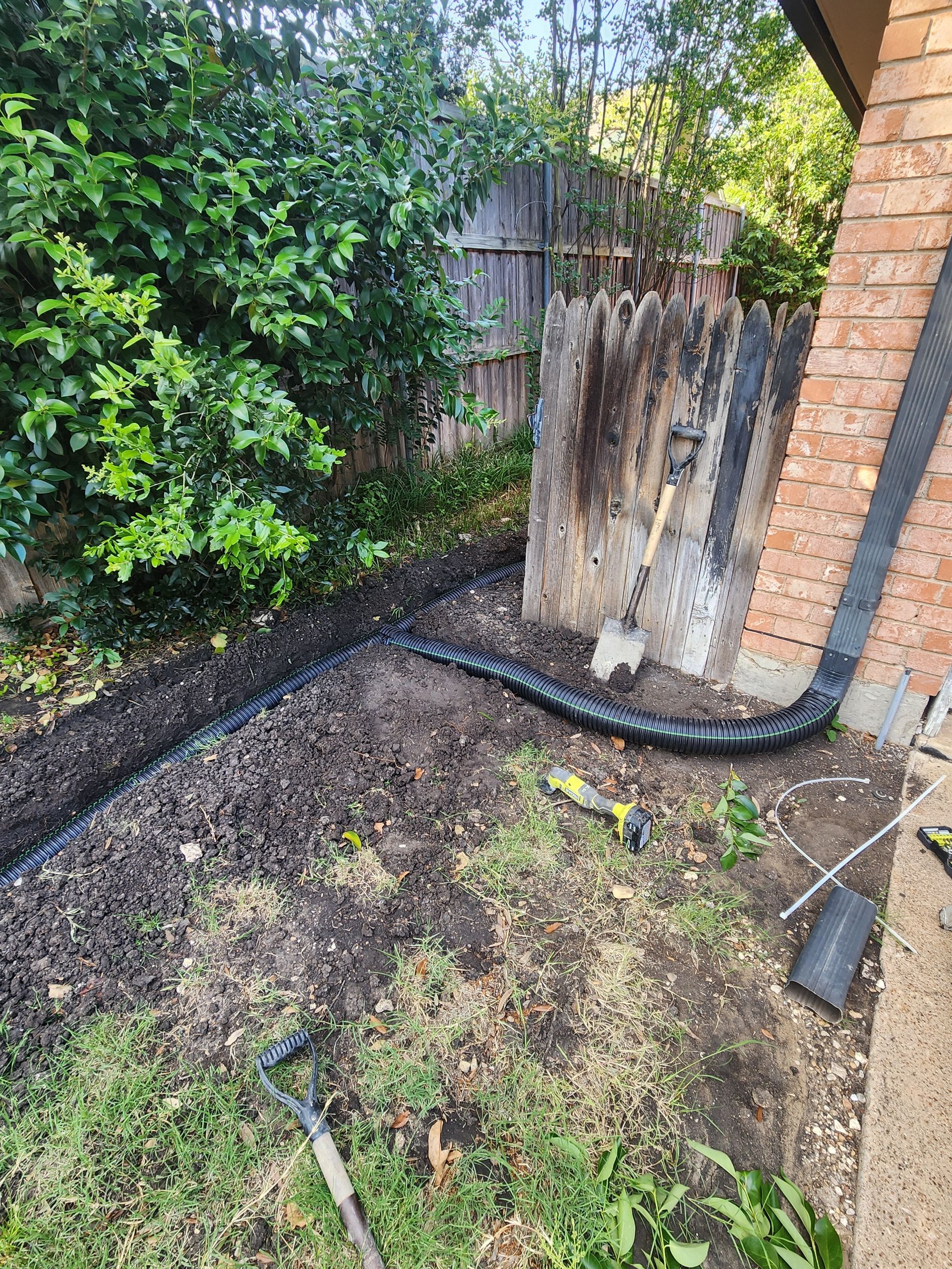 Drainage pipe installation in a backyard garden, along a fence and building, with tools and mulch.