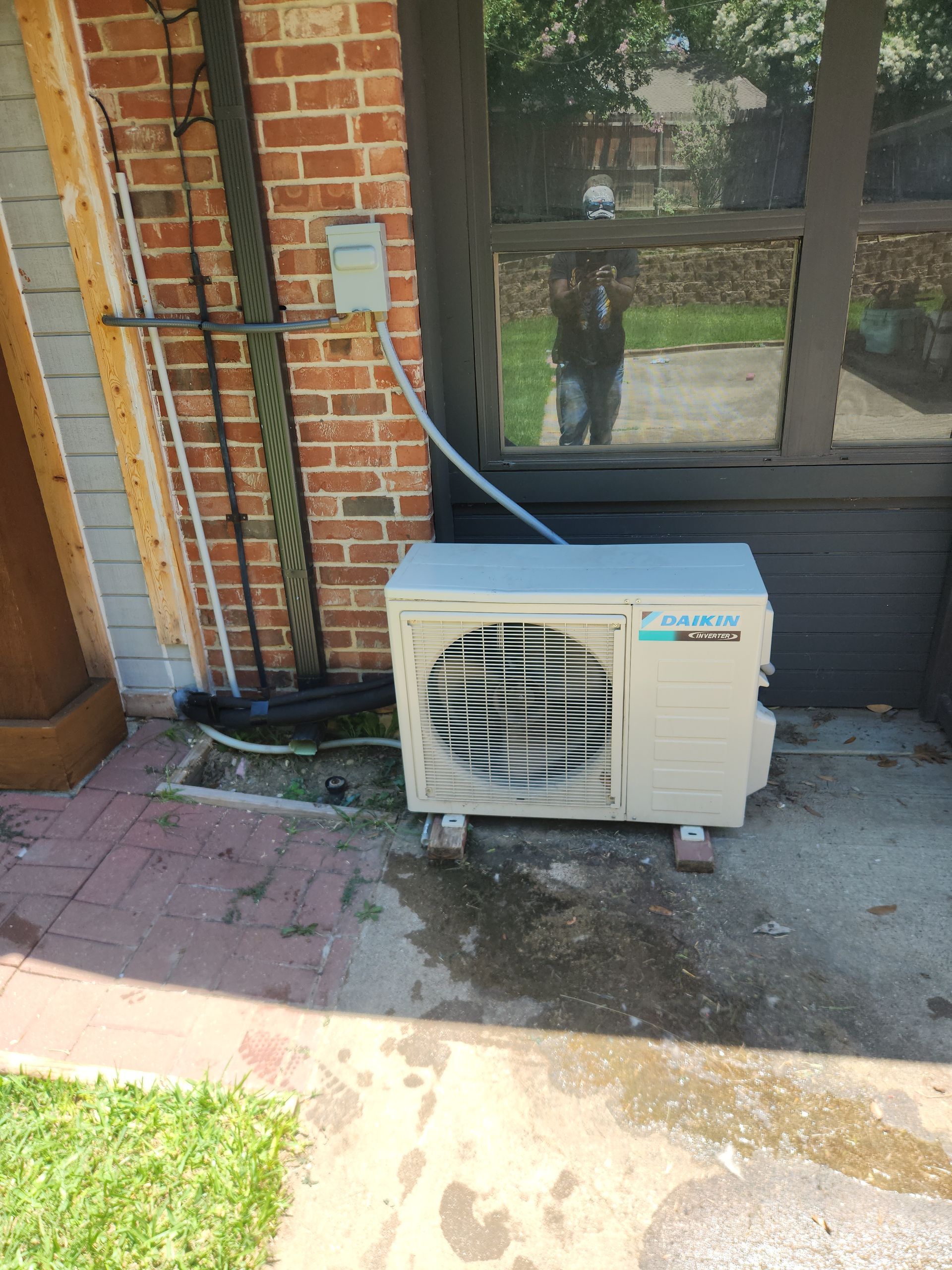 An outdoor Daikin air conditioning unit next to a brick wall and window, sitting on concrete blocks.