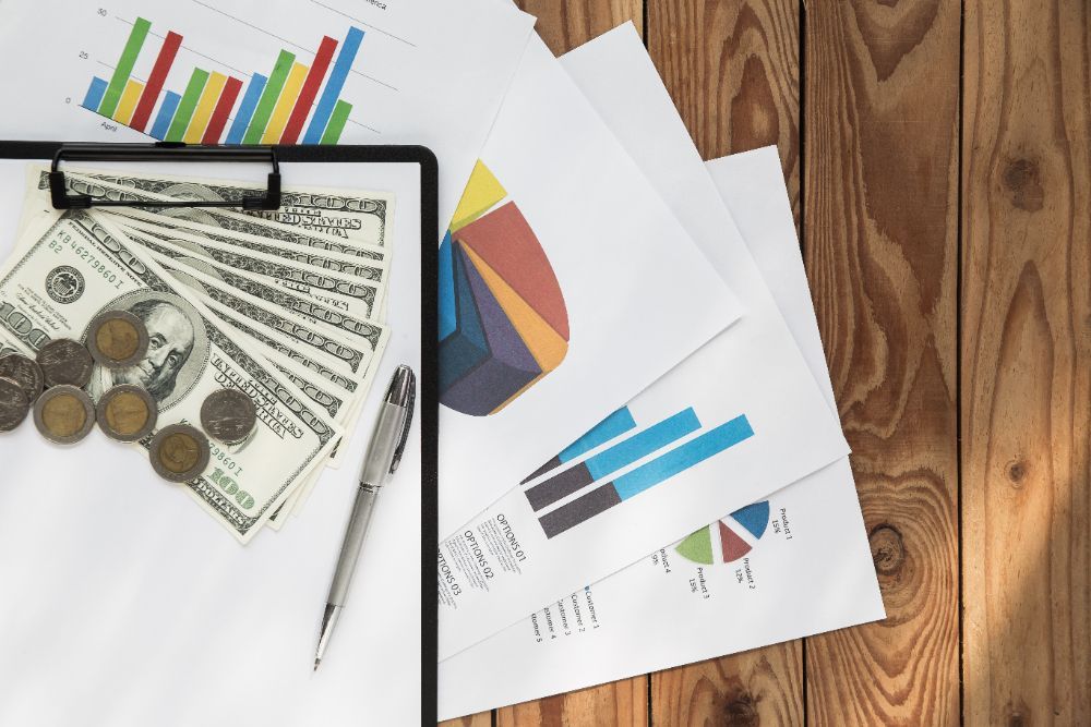 A clipboard with money, coins, graphs, and a pen on a wooden table.