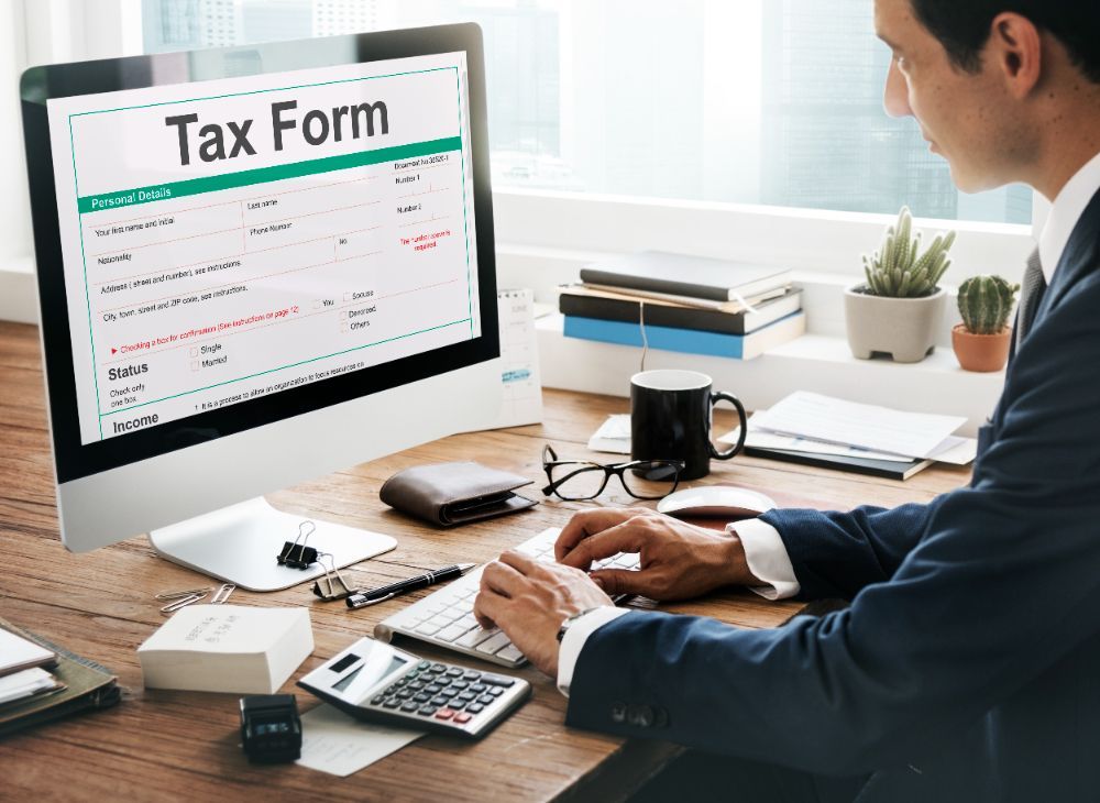 A man is sitting at a desk using a computer to fill out a tax form.