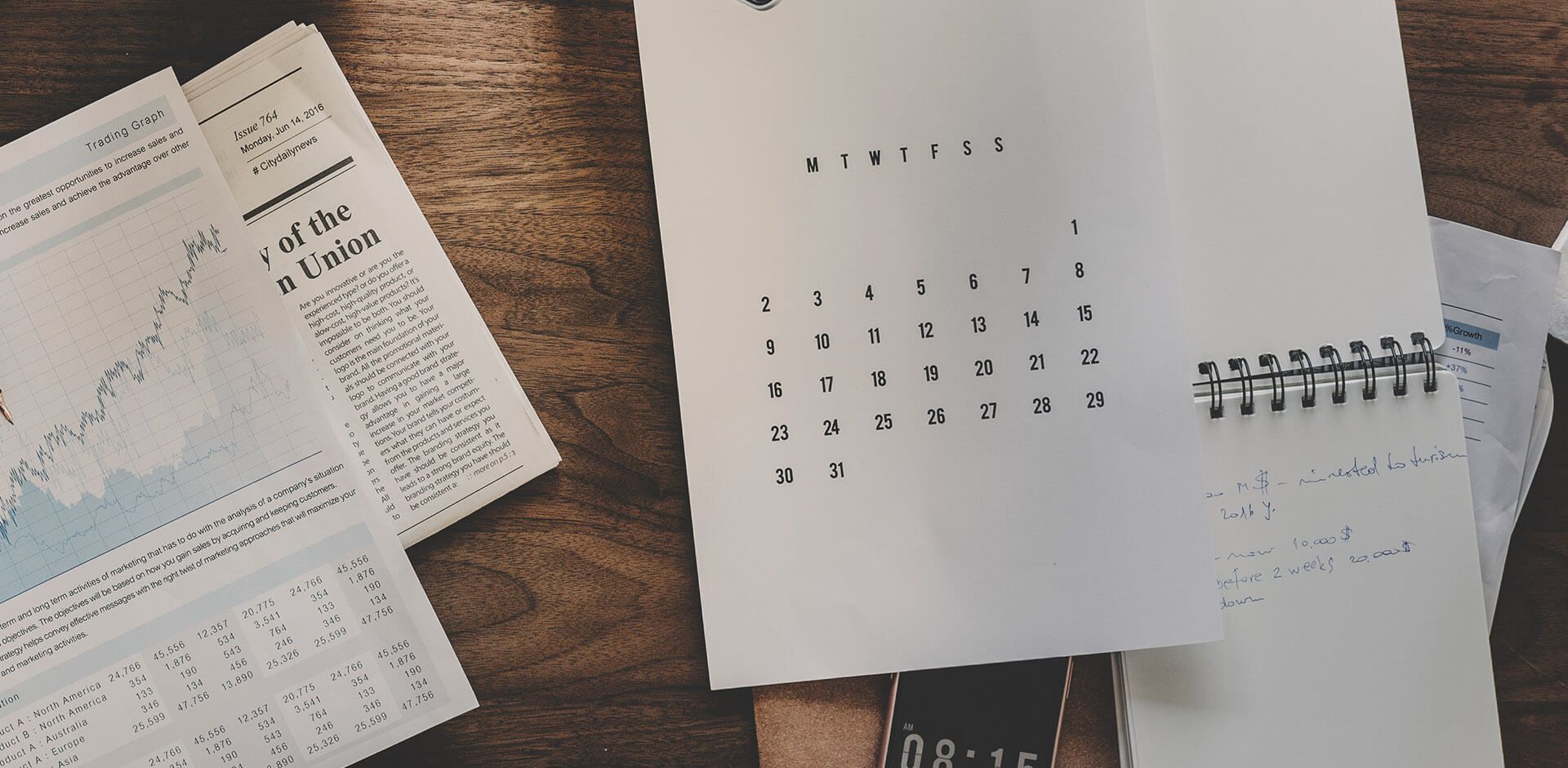 A calendar is sitting on a wooden table next to a newspaper and a watch.