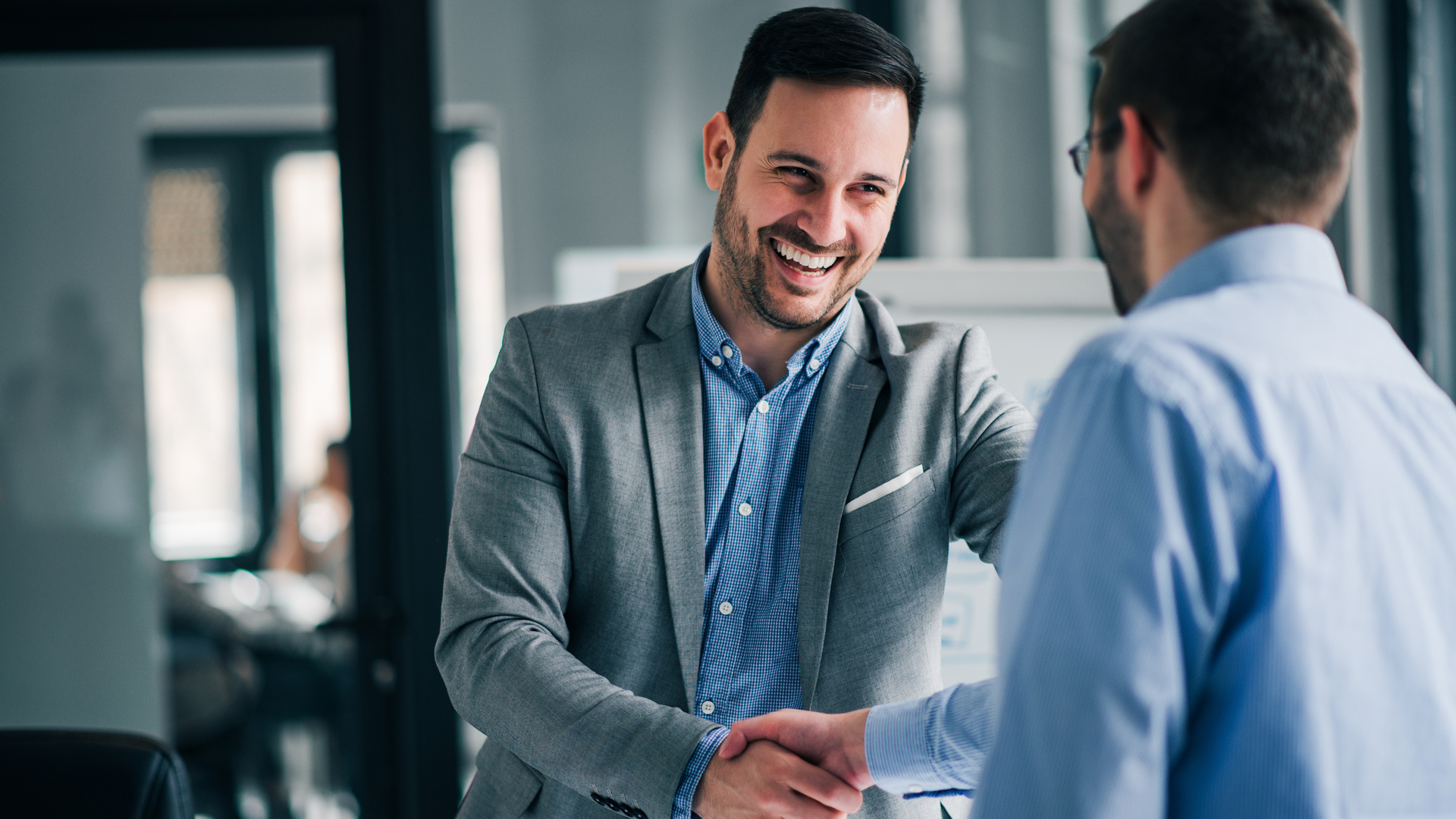 Two men are shaking hands in an office and smiling.