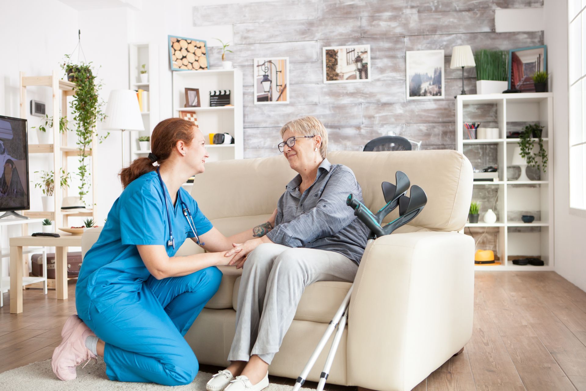 A nurse is kneeling down next to an elderly woman sitting on a couch.