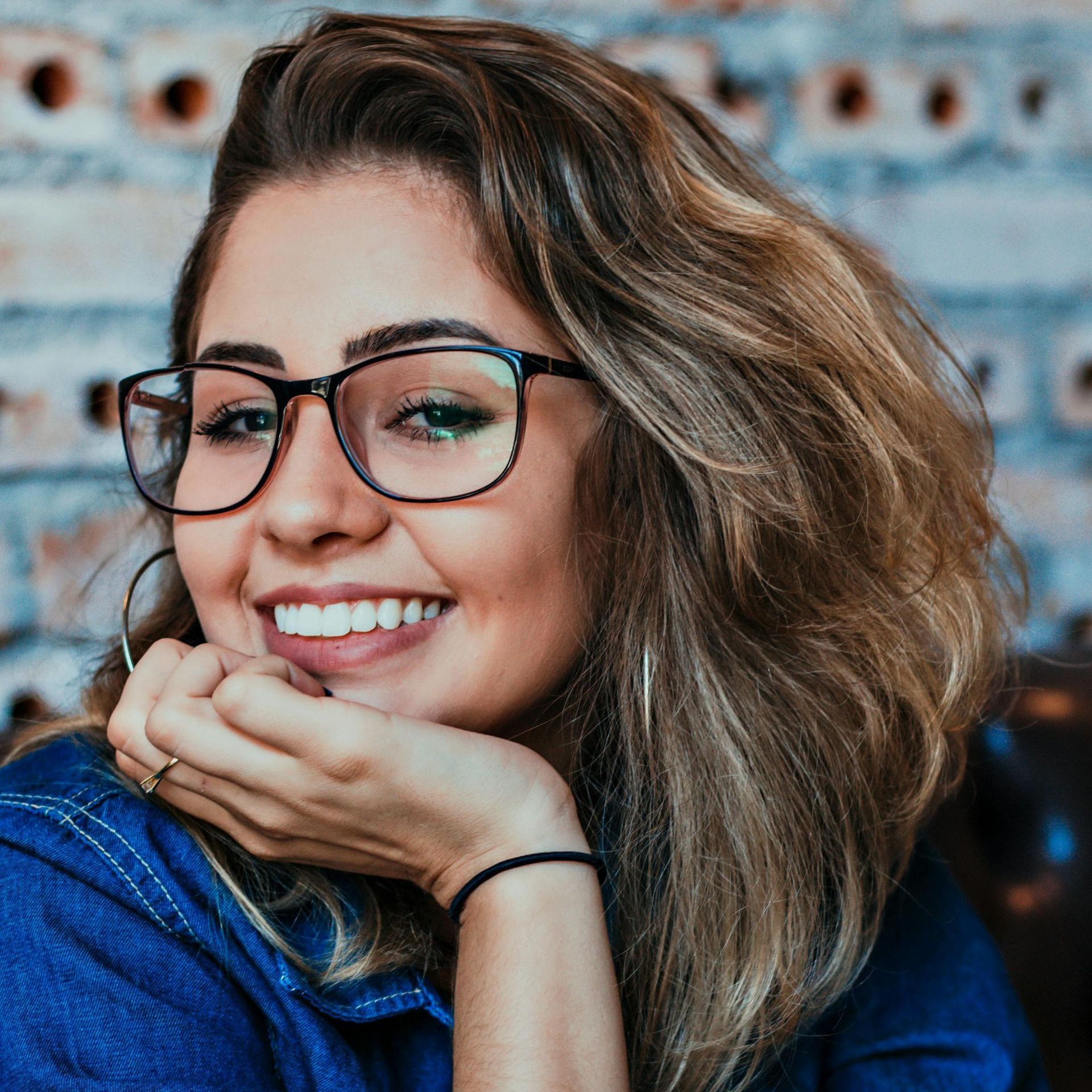 A woman wearing glasses is smiling with her hand on her chin.