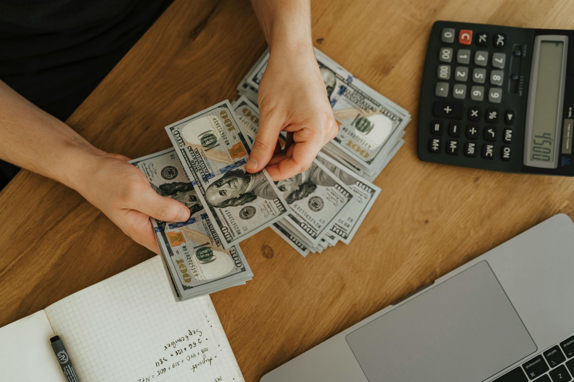 A person is counting money on a table next to a laptop and a calculator.