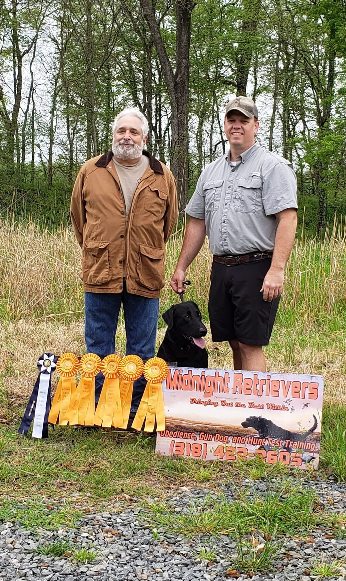 Two men and a black dog pose with ribbons and a sign outdoors.