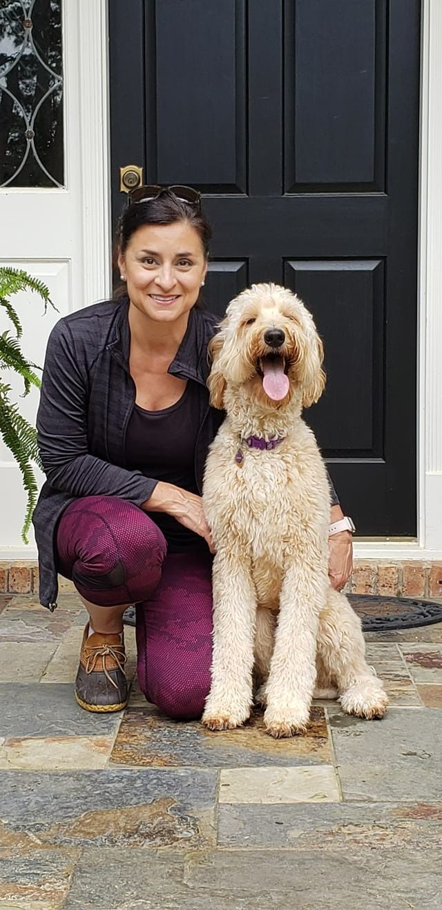 Woman with a light brown dog in front of a black door. The dog is smiling. The woman wears purple pants.