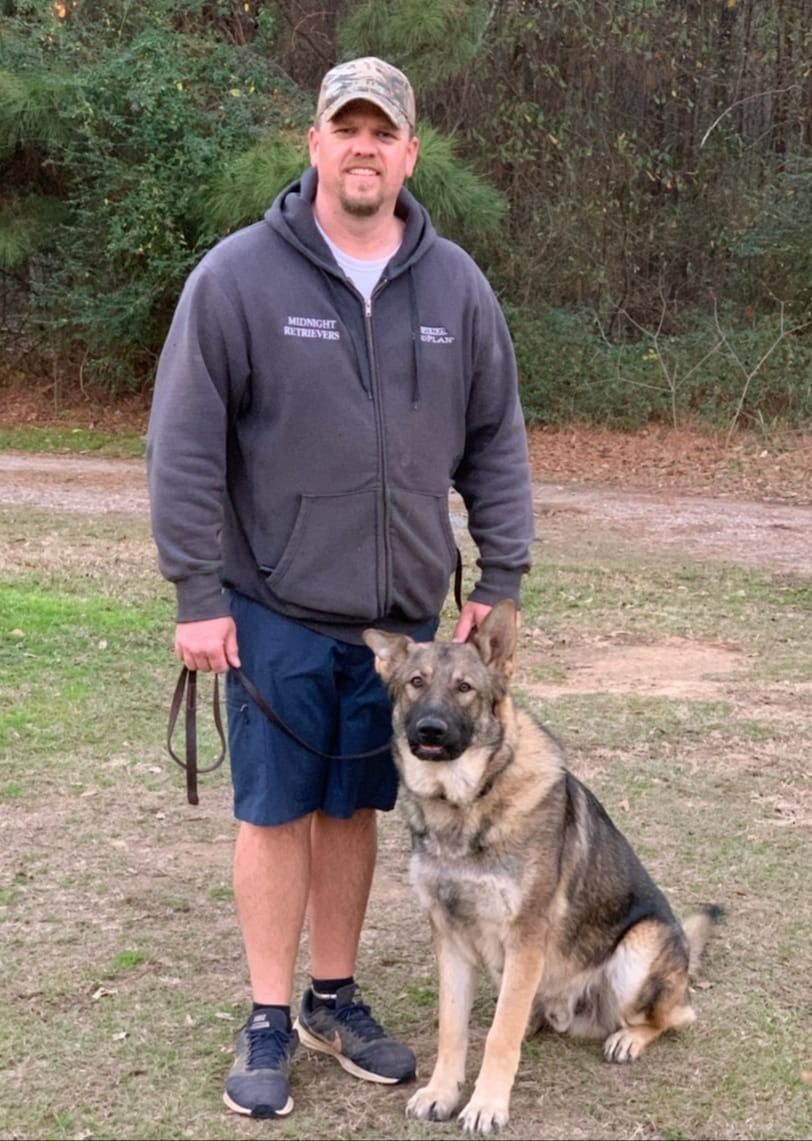 Man in gray hoodie and cap with a tan and gray dog, standing in a grassy area.