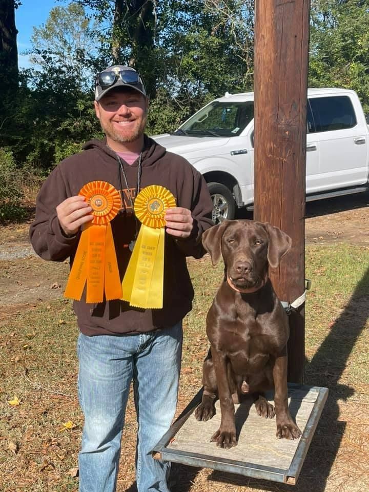 Man holds ribbons, smiling next to a brown dog sitting on a platform. A white truck is in the background.