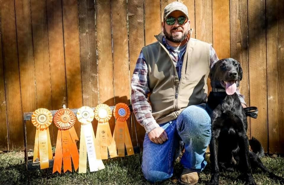 Man kneeling next to a black dog, with ribbons hanging on a wooden fence.