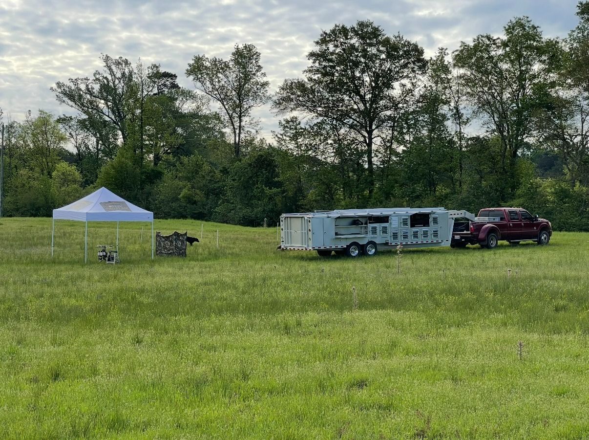 A white trailer and truck parked in a grassy field with trees in the background. A tent is set up nearby.