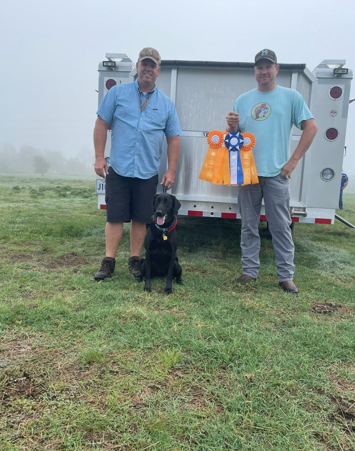Two men and a black dog stand outdoors with a ribbon. The dog sits in front of a man holding a ribbon.