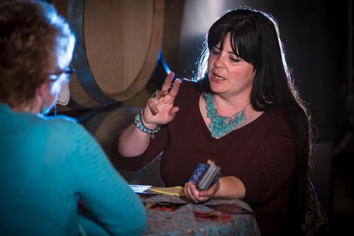 A woman is sitting at a table talking to another woman while holding a tarot card.