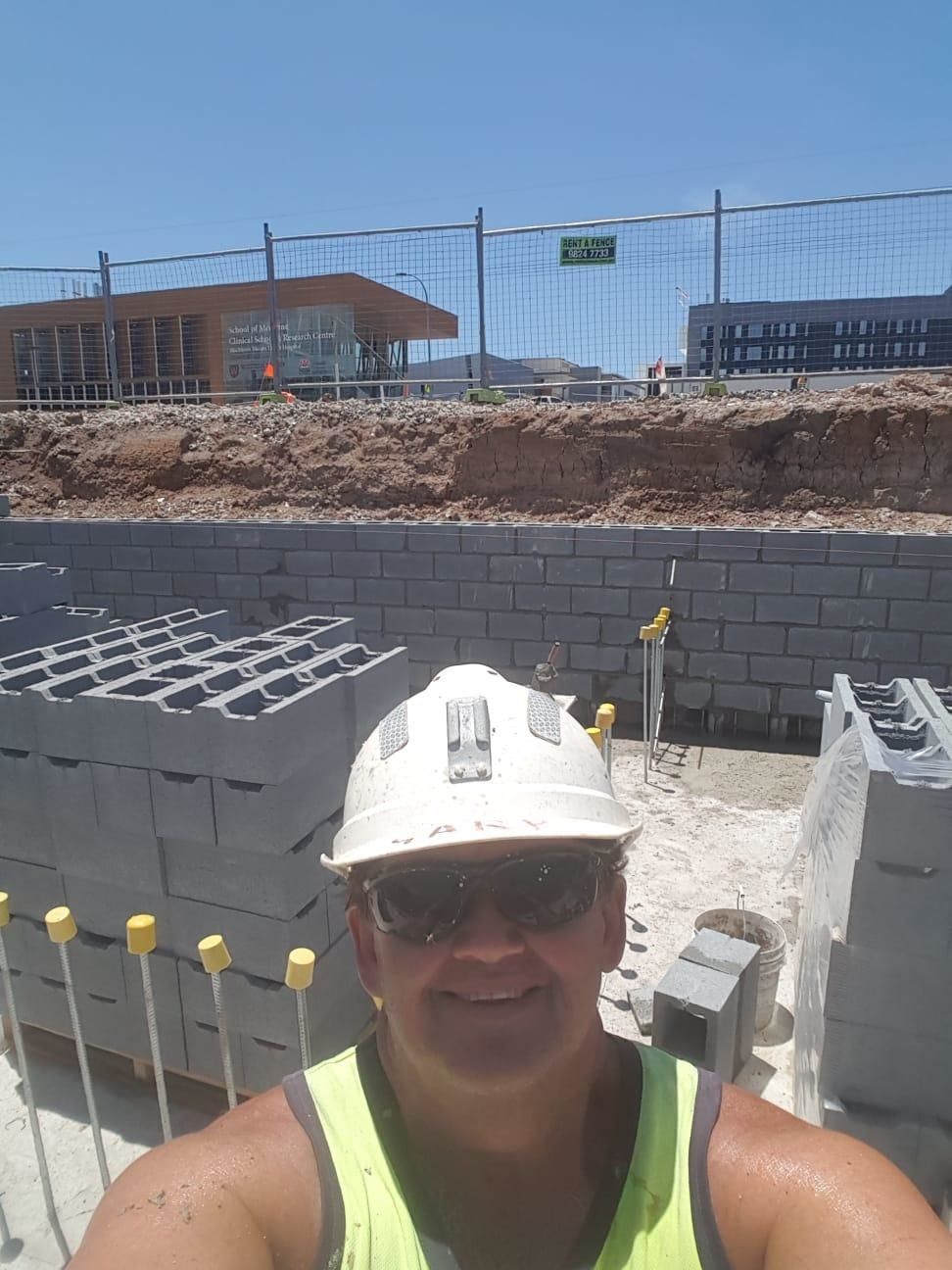 Person in construction site wearing hardhat, sunglasses, and safety vest, smiling. Blocks of concrete in the foreground.