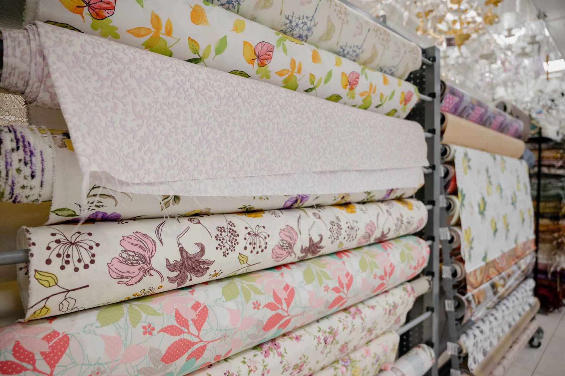 Shelves stacked with rolls of colorful floral fabric in a store. Various patterns and hues, including pink, green, and white are visible.
