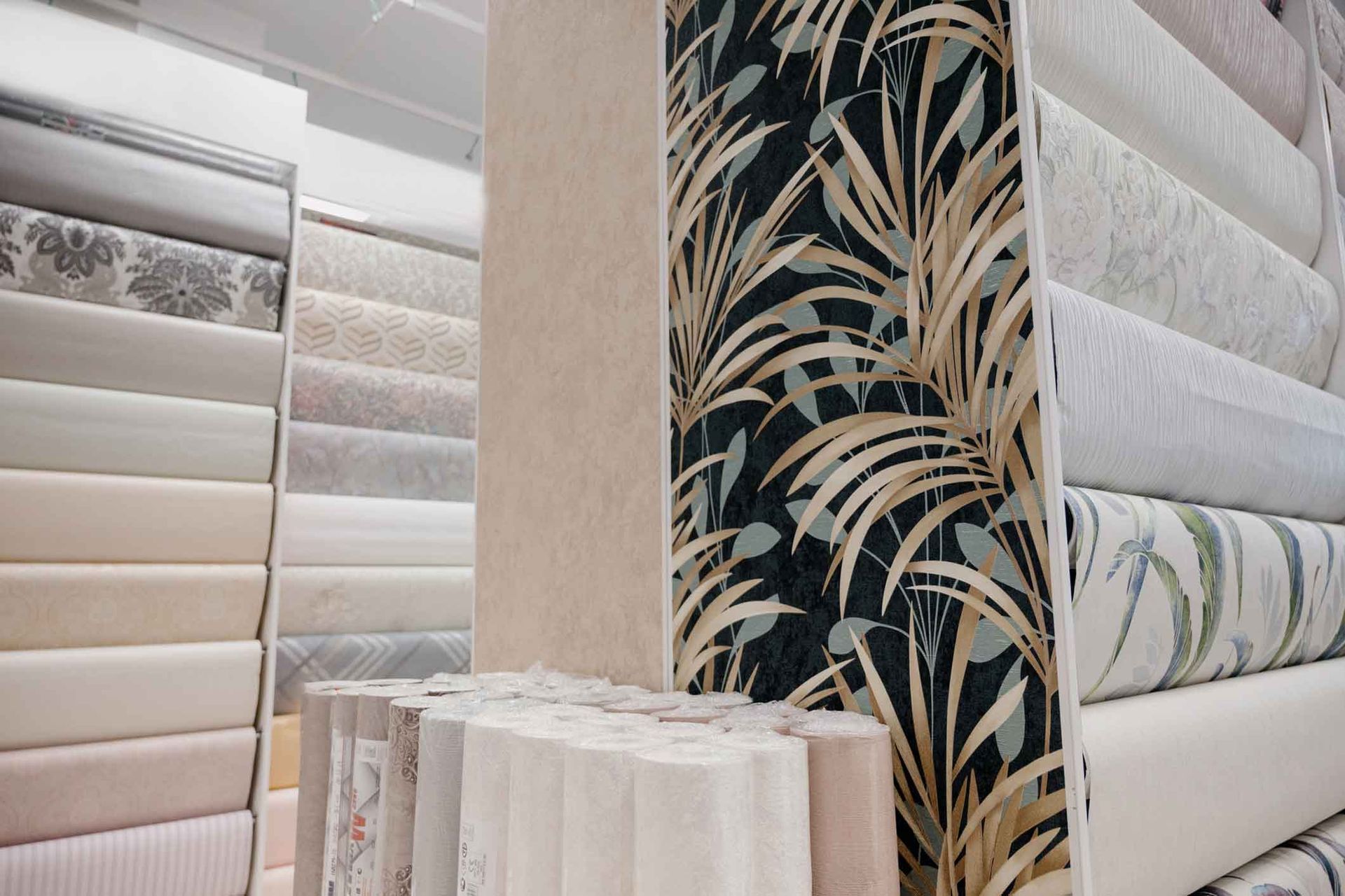 Wallpaper display in a store, showcasing various rolls of paper with different patterns and textures. Focus on a dark leafy design.