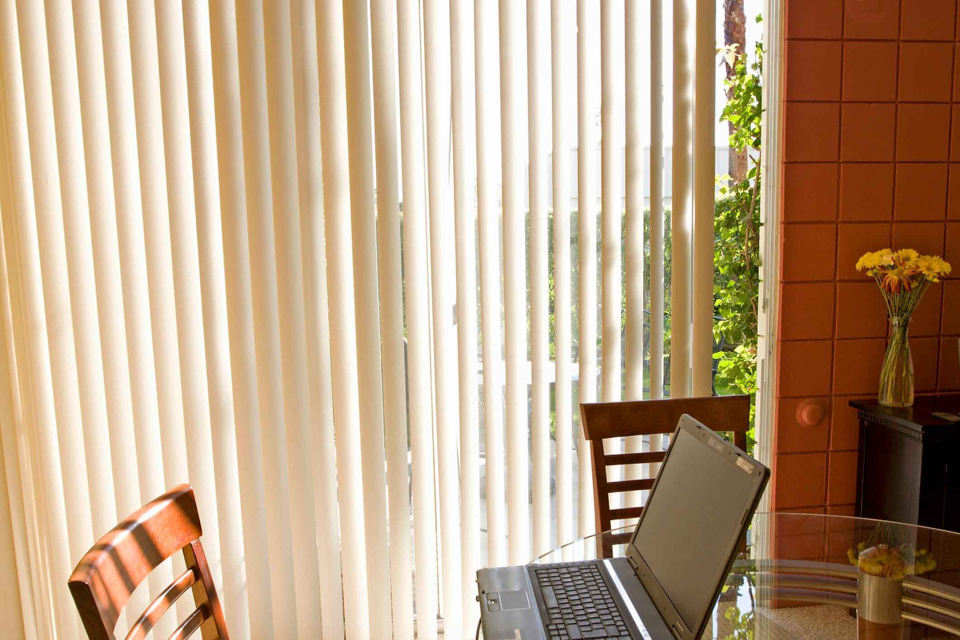Laptop on table near a window with vertical blinds. Chair in foreground, vase of flowers on side table.