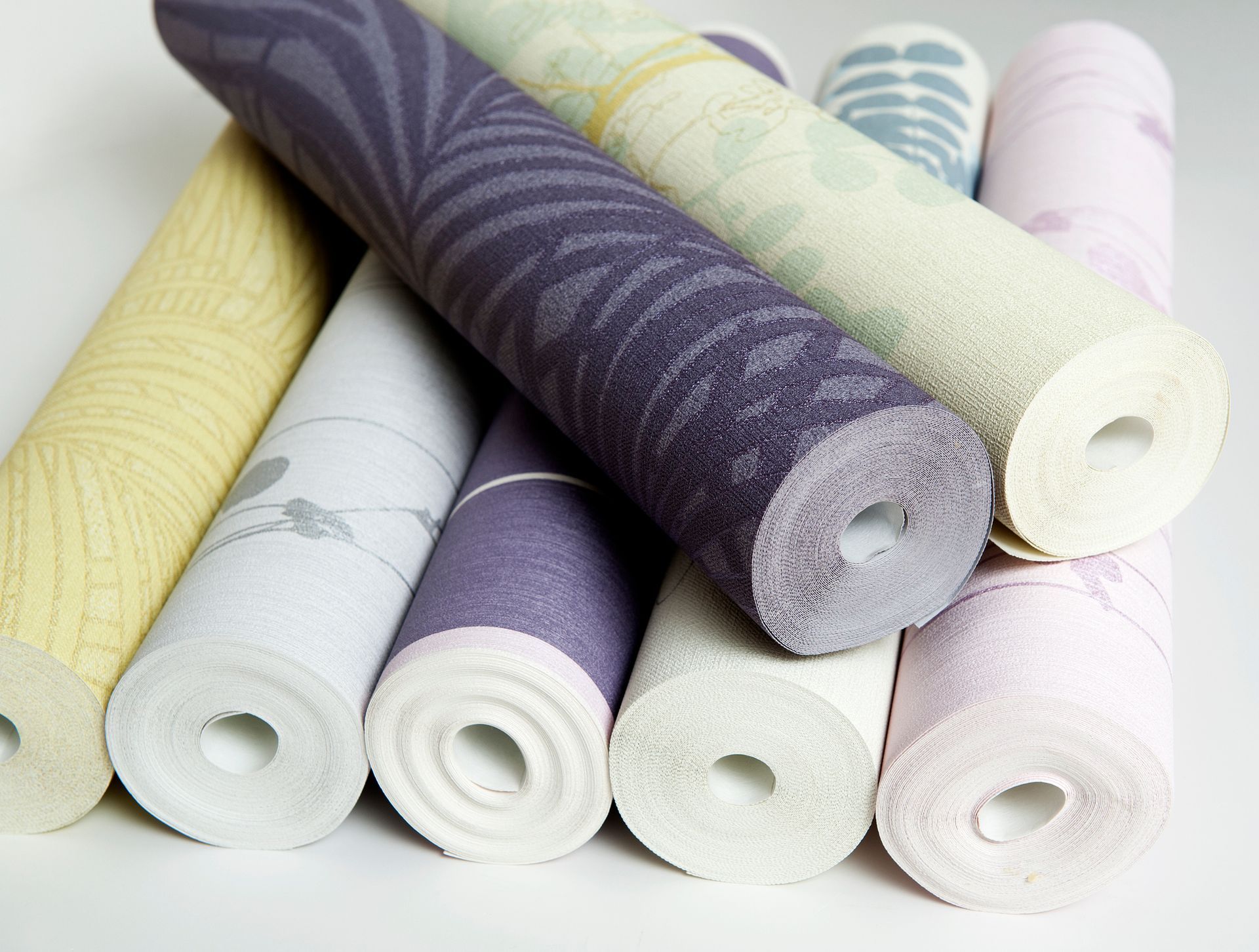 Close-up of several rolls of patterned wallpaper in various colors, stacked on a white surface.