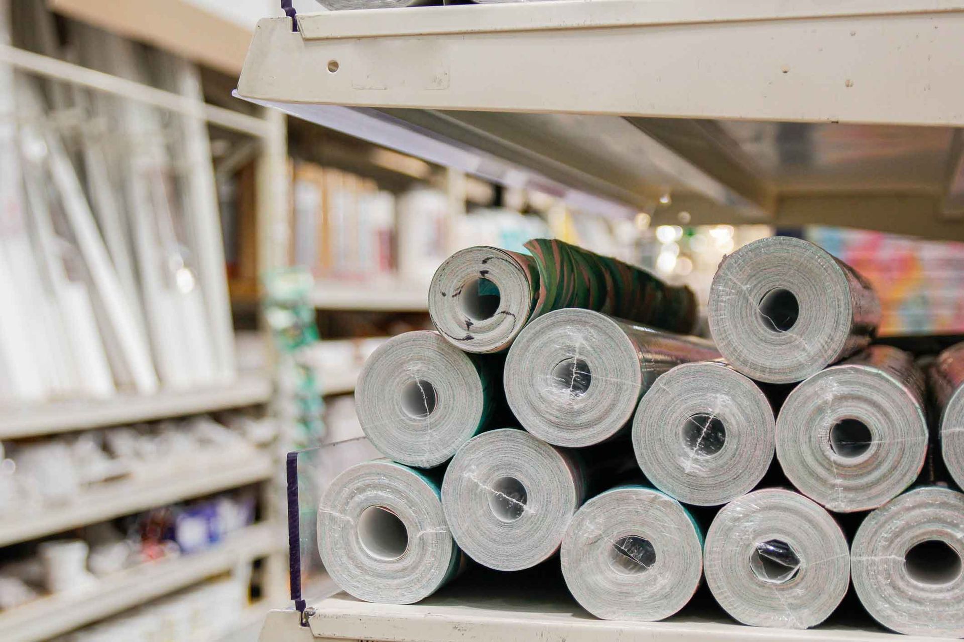 Rolls of patterned wallpaper stacked on a shelf in a store, with other building materials visible in the background.