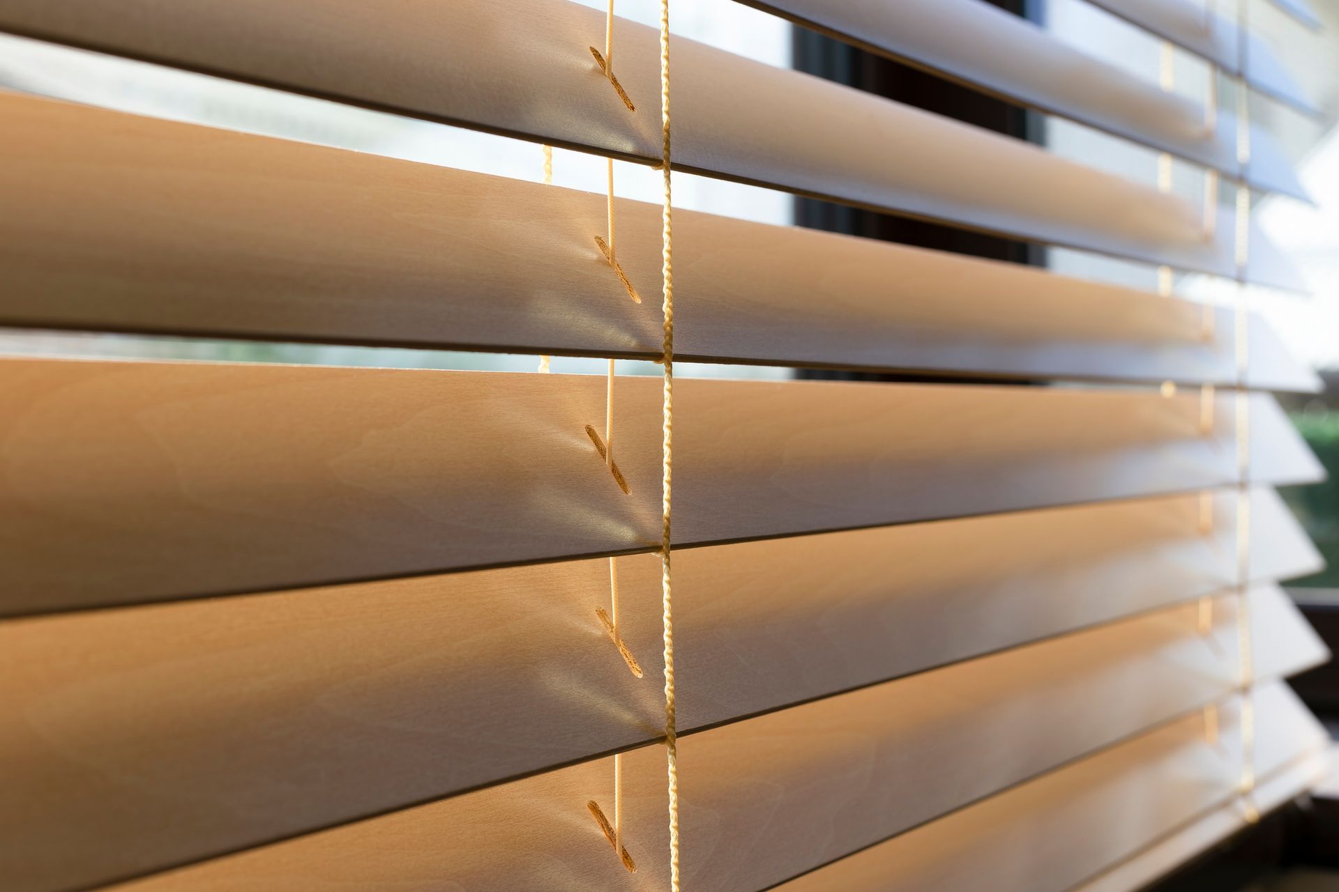 Wooden venetian blinds, angled to filter light from a window. Beige slats are connected by thin cords.