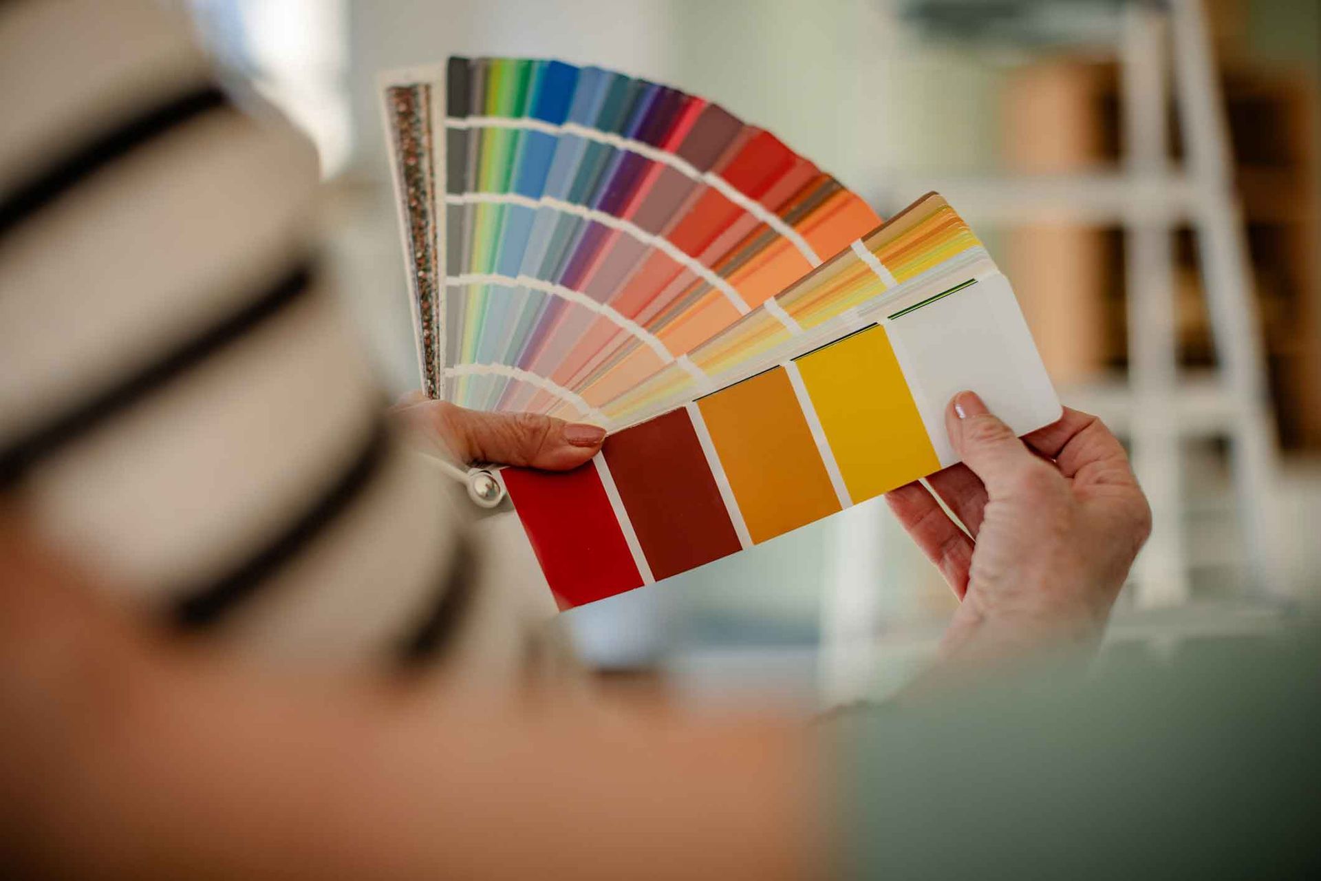 Person holding a color palette fan, selecting paint colors, with a partially visible striped shirt and a ladder in the background.