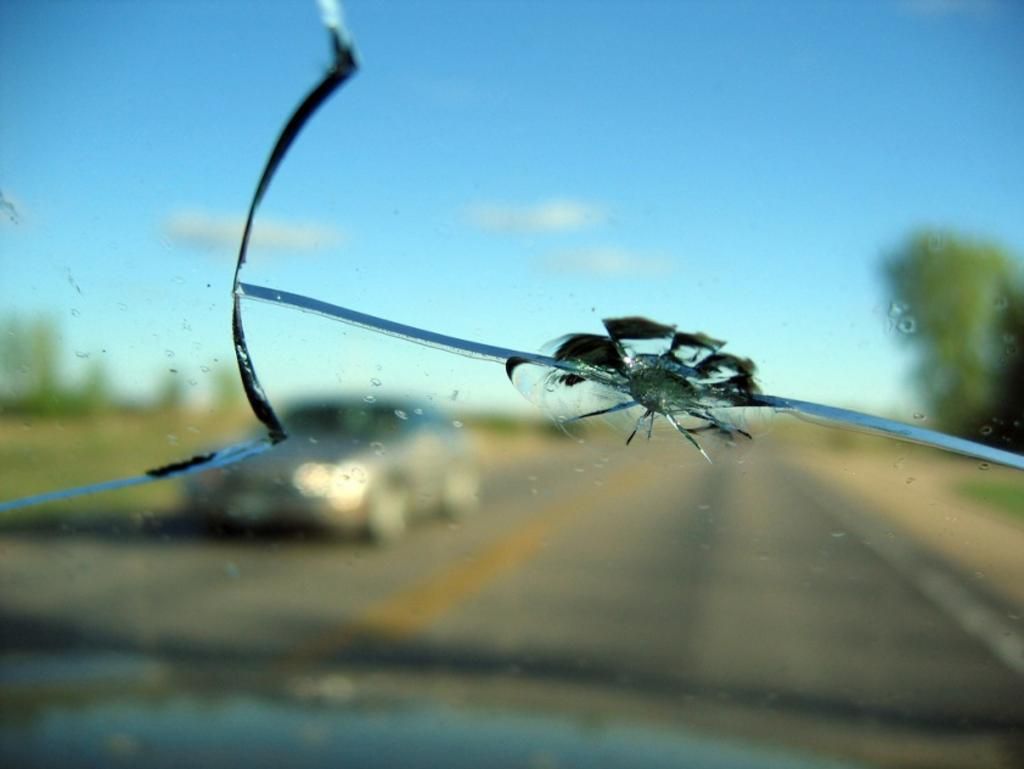 A close up of a broken windshield with a car in the background.