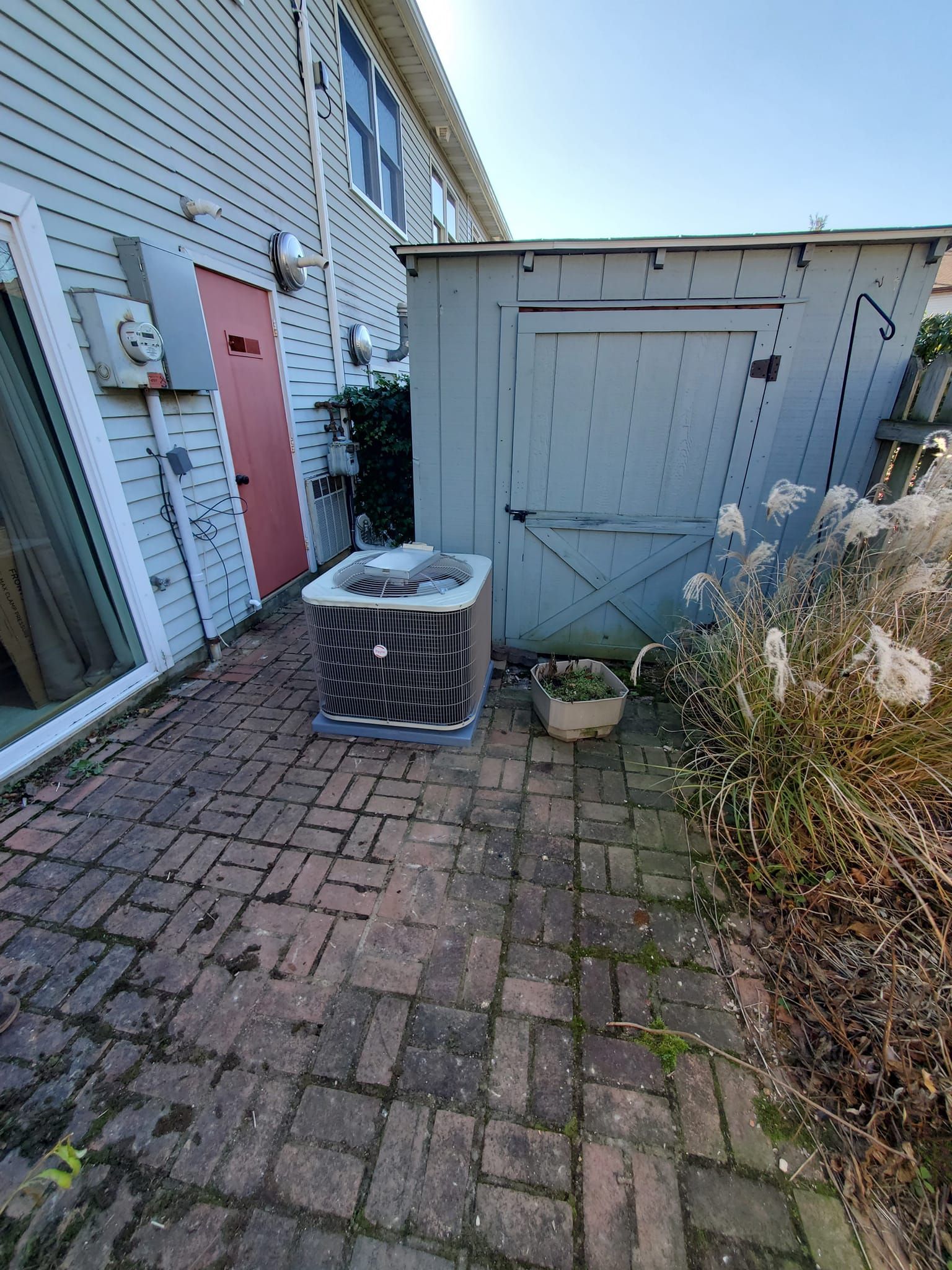 An air conditioner is sitting on a brick patio next to a shed.