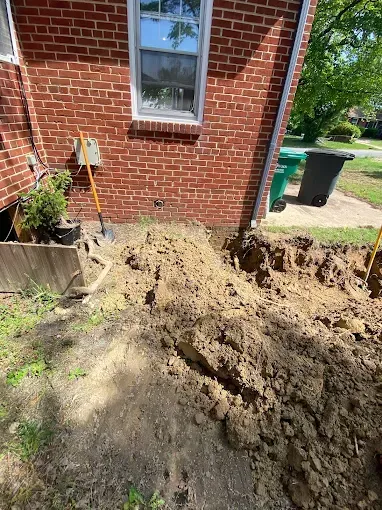 Brick house exterior with a trench dug along the foundation, pile of dirt in the foreground.