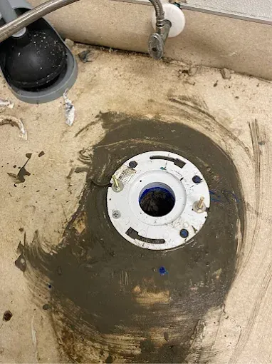 A white toilet flange with four bolts, surrounded by grey sealant, on a concrete floor.