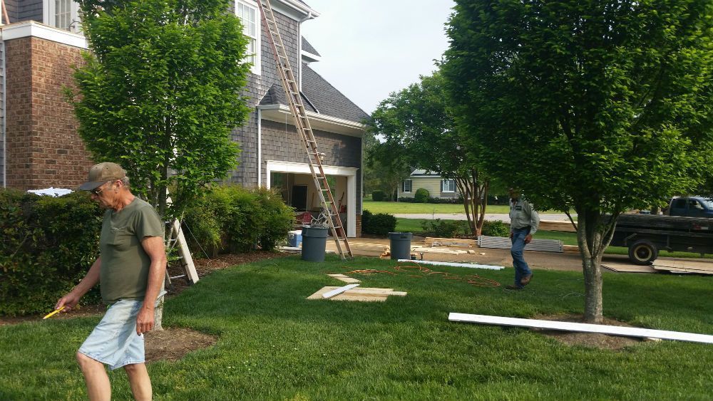 Two men working on a house exterior. One measures trim, the other near a ladder. Green grass and trees surround.