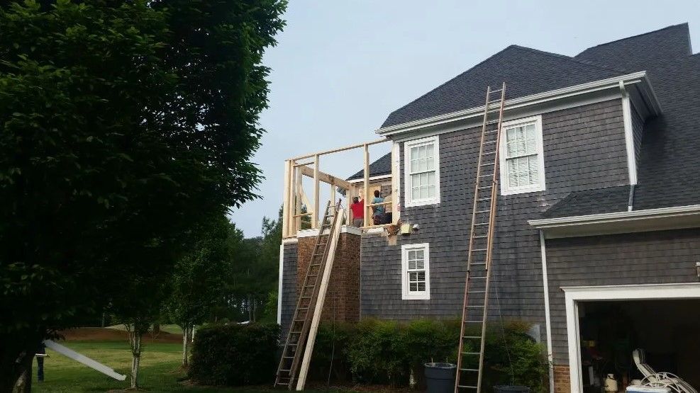 Construction of a wooden deck extension on a two-story gray house. Two ladders lean against the house.