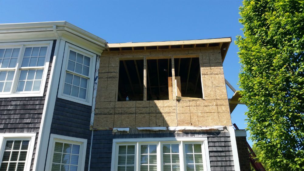 Construction of a second-story addition on a blue-shingled house. Window openings visible; plywood siding.