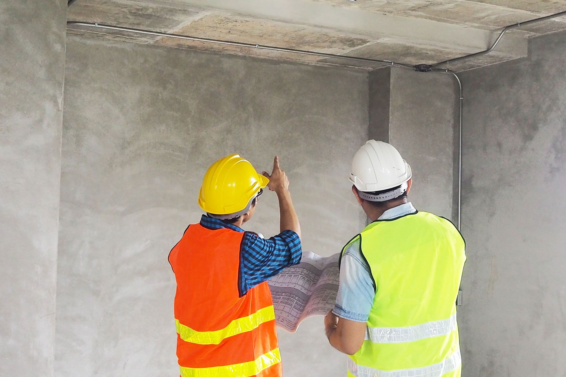 Two construction workers in safety vests and hard hats reviewing blueprints inside a building.