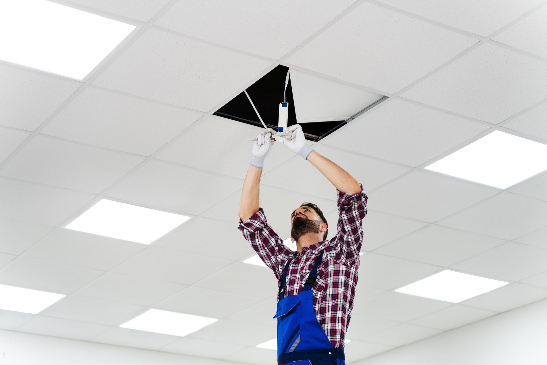 Person in blue overalls on a ladder, working on a ceiling panel. The setting is an office with overhead lighting.