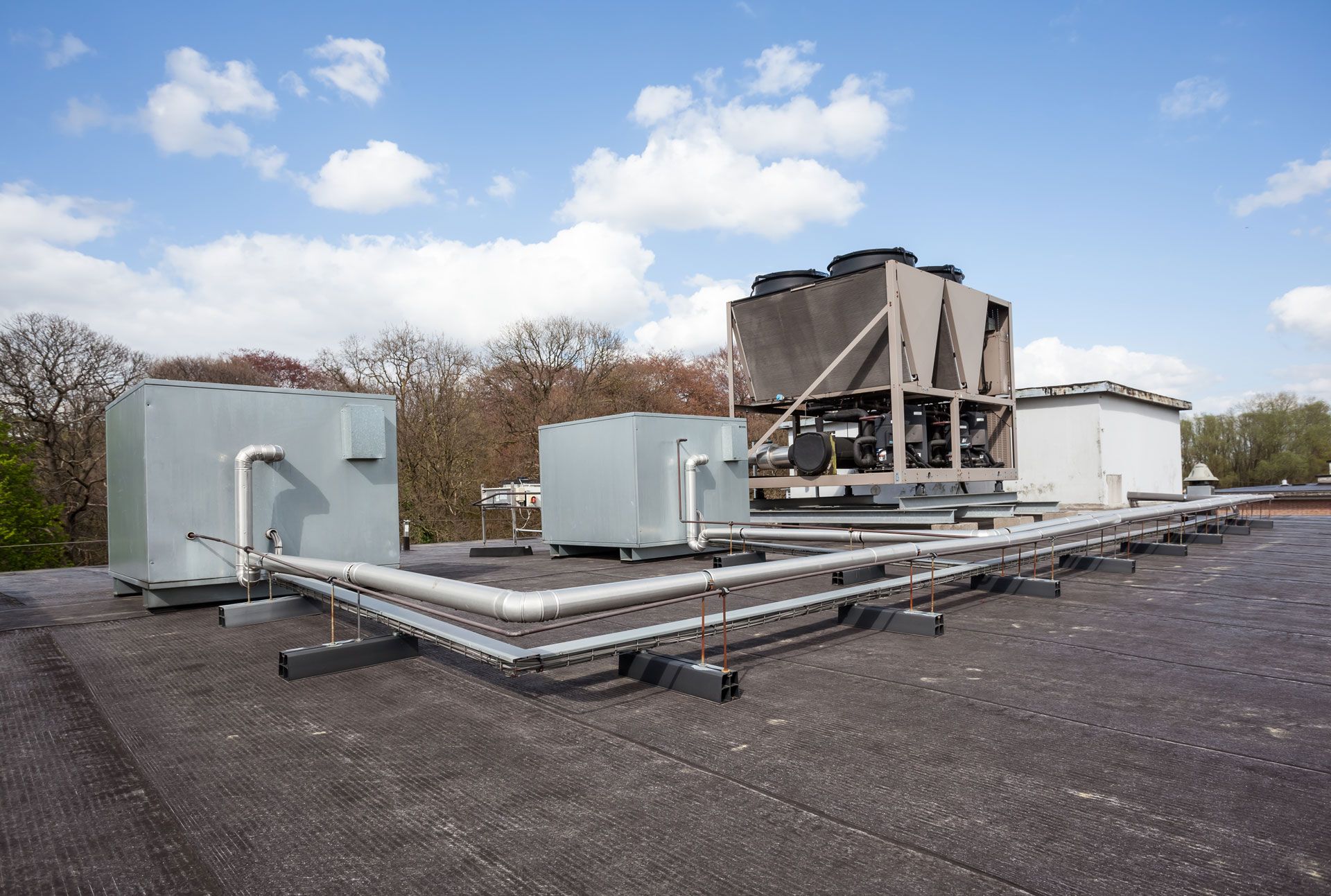 Rooftop HVAC units against a cloudy sky. Gray metal equipment, pipes, and supports on a flat, dark roof.