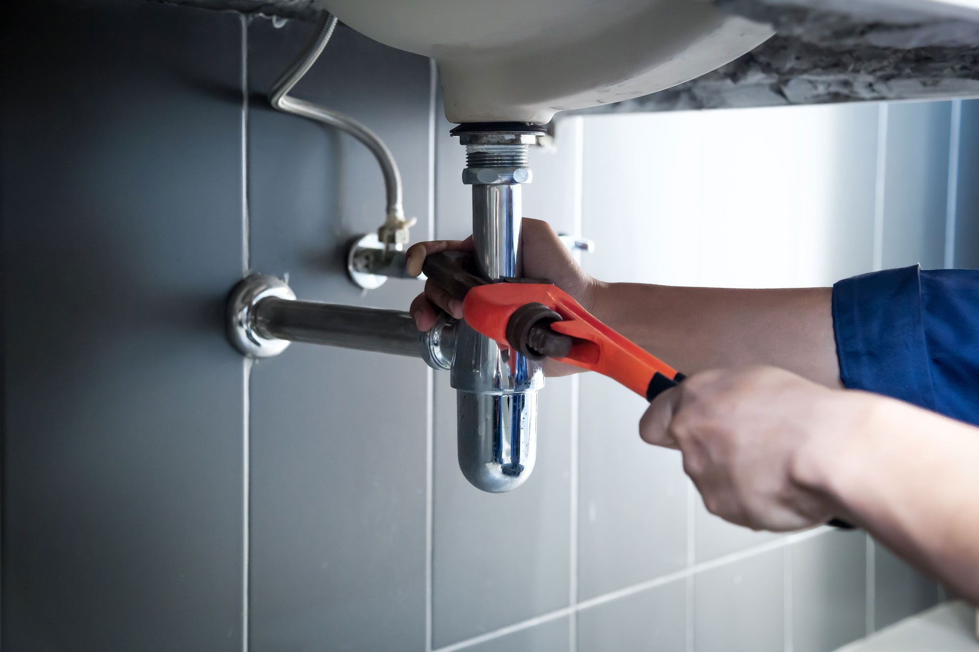 Plumber using a wrench to work on a sink's drain pipes. Silver pipes, blue shirt, gray tiles.