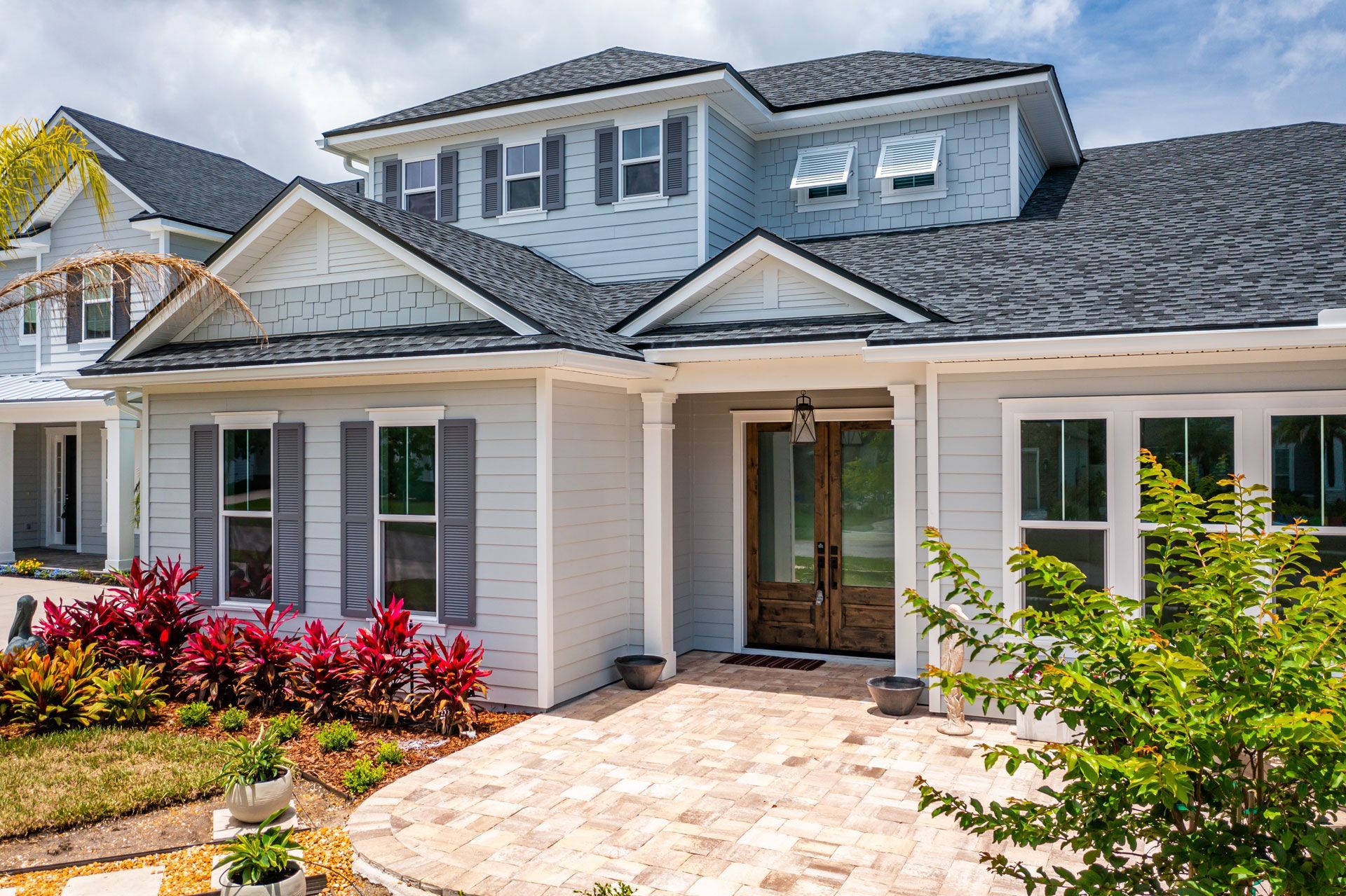 Light blue two-story house with dark gray roof, red and green landscaping, and brick walkway.