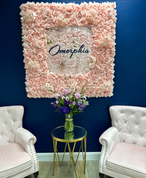 Omorphia Aesthetics Front Lobby is a room with two chairs , a table , and a wall of pink flowers behind their sign.
