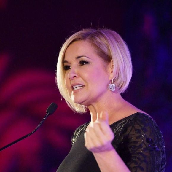 Sandra Peebles with blonde bob haircut speaking at a podium, gesturing with a raised thumb.