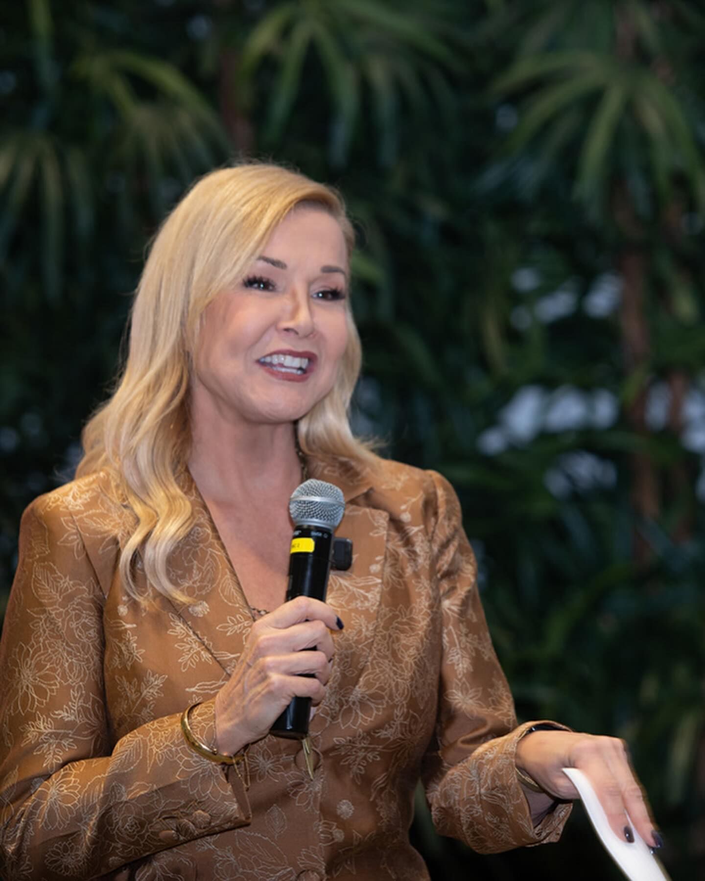 Sandra Peebles speaking into a microphone, wearing a gold blazer, smiling.