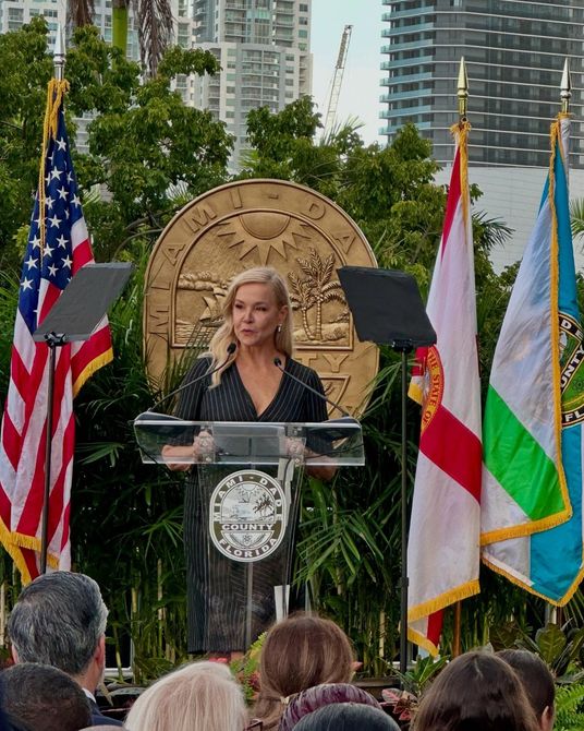 Sandra Peebles speaking at a podium with flags in front of a city seal. 