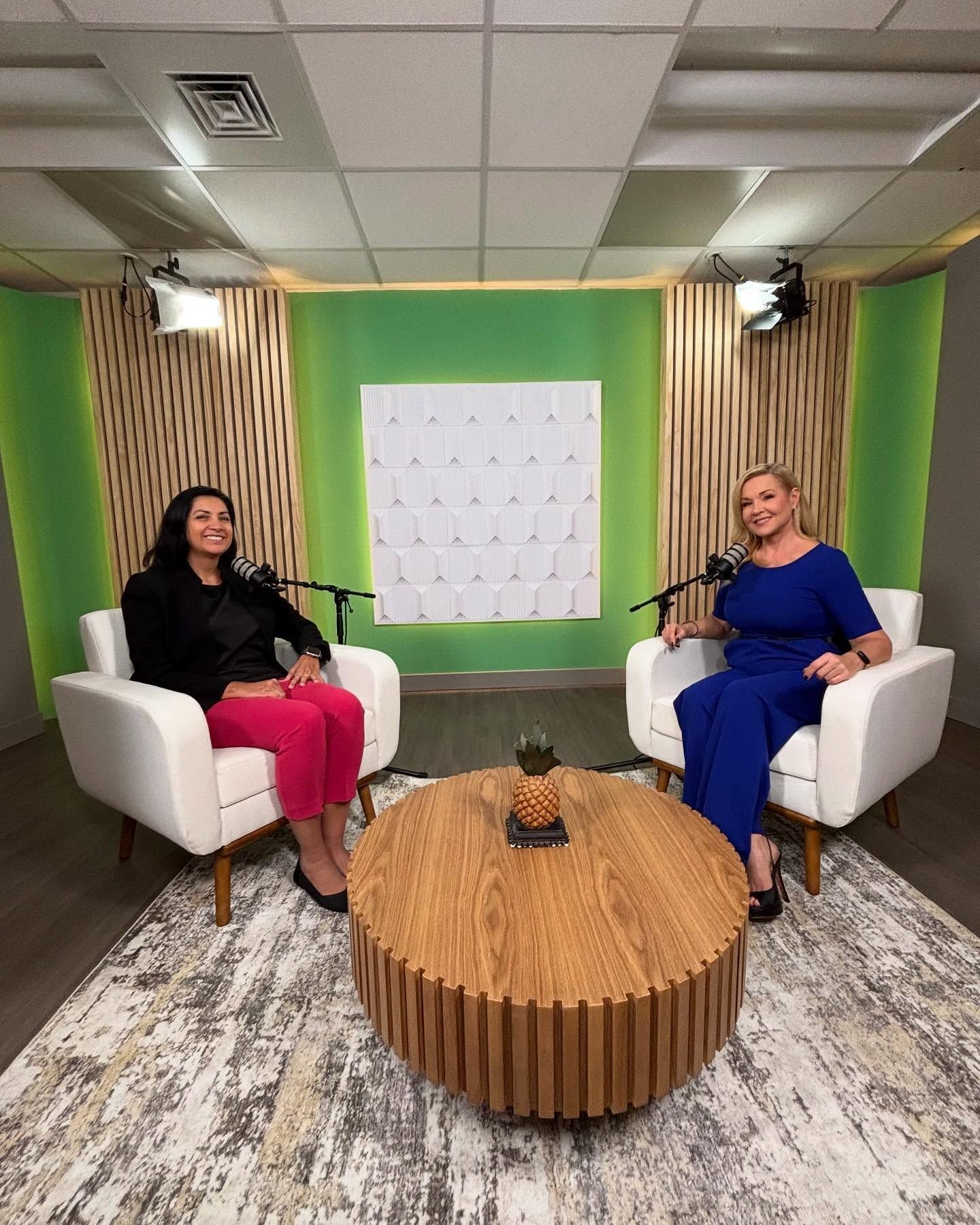 Two women seated in a podcast studio with a wood and green backdrop, microphones, and a round coffee table.