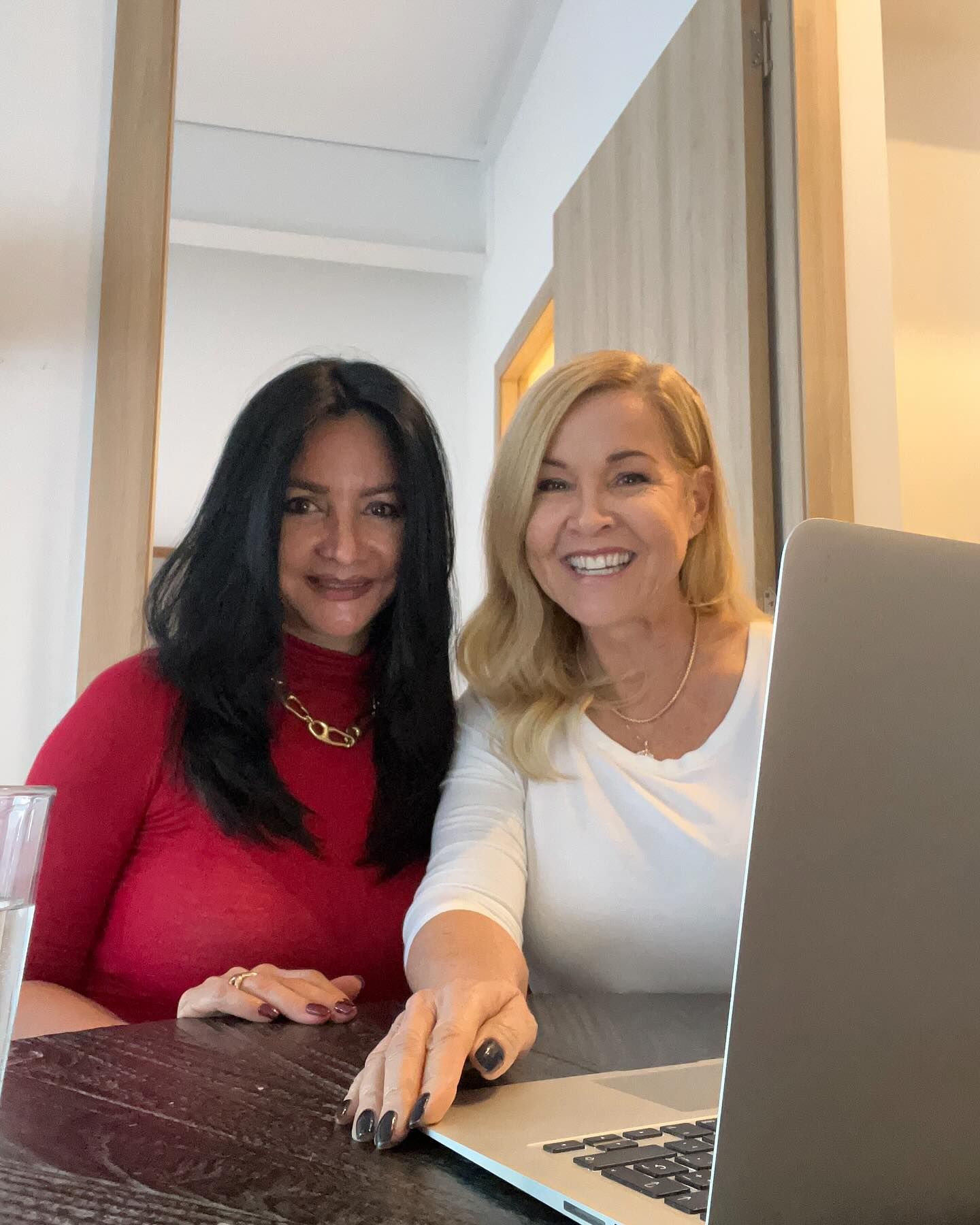 Two smiling women seated at a table, one in red, one in white. 