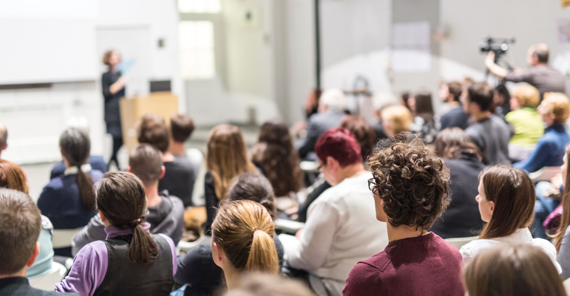 A presenter speaks to an audience in a lecture hall. People are seated and looking forward.