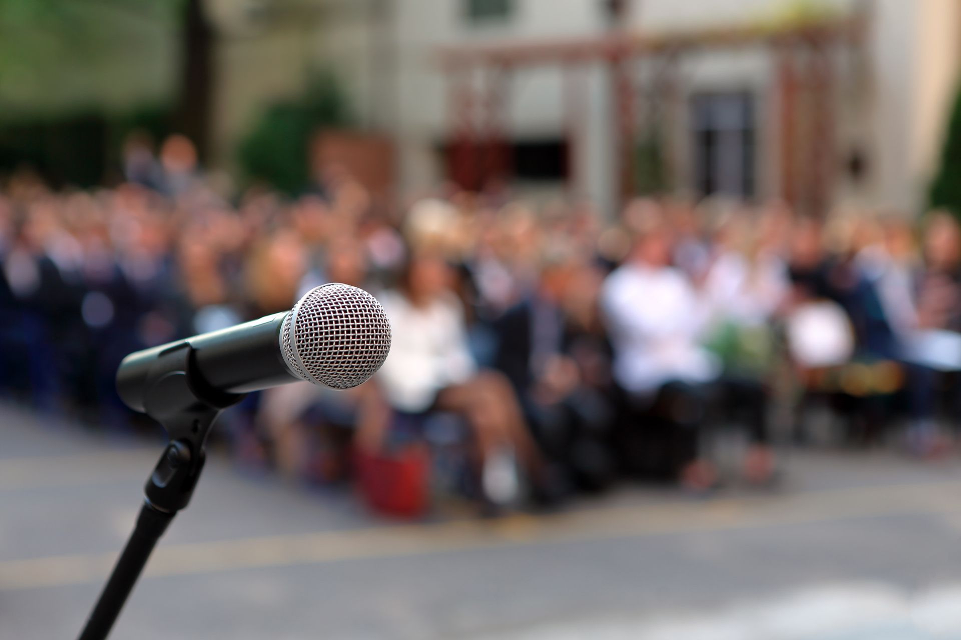 Microphone on stand, blurred audience seated outdoors, possibly at an event.