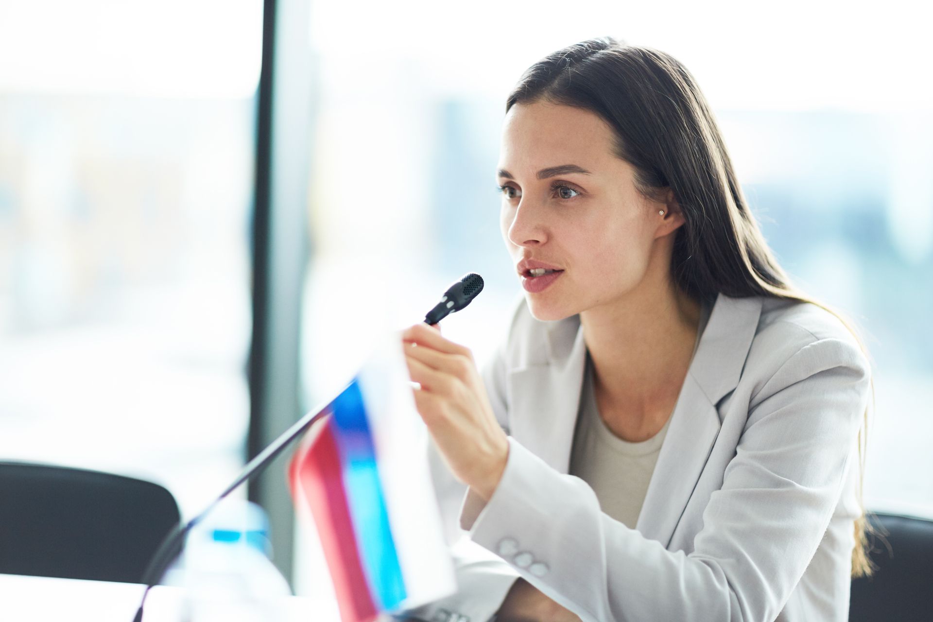 Woman speaking into a microphone, holding a flag with red, blue, and white stripes, in a bright room.