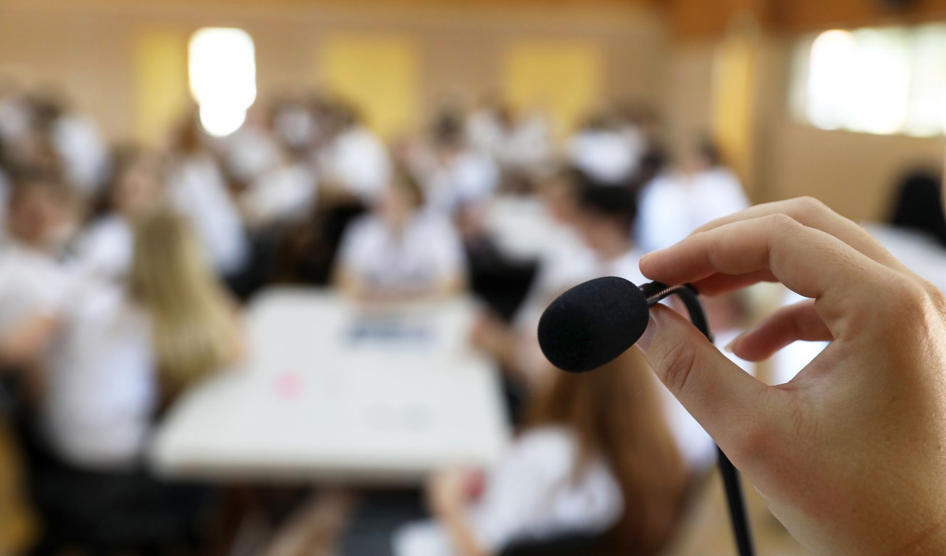 Hand holding a small microphone in front of a blurry classroom scene with people seated at tables.