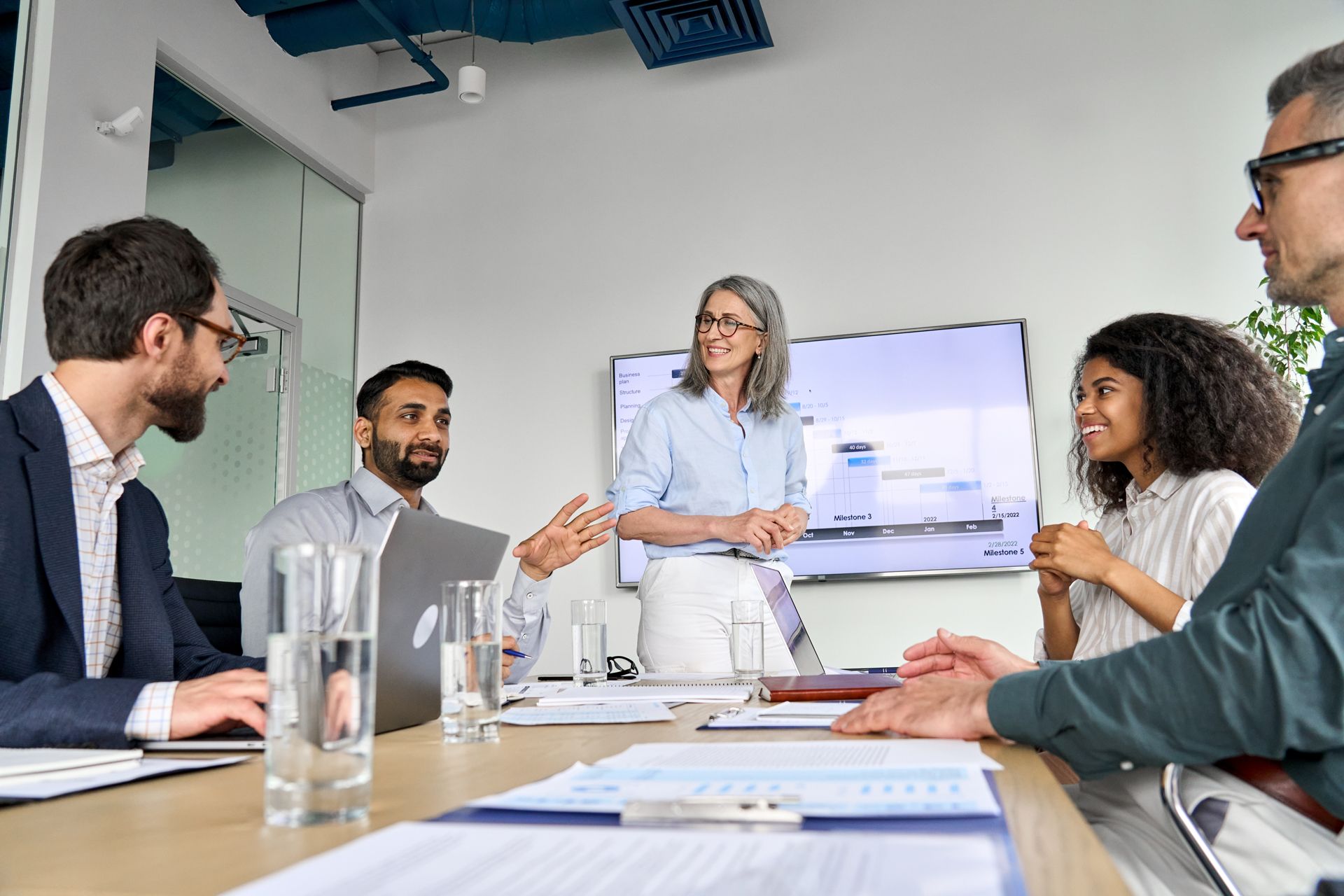 Business team in a meeting around a table, one person presenting, whiteboard in the background.